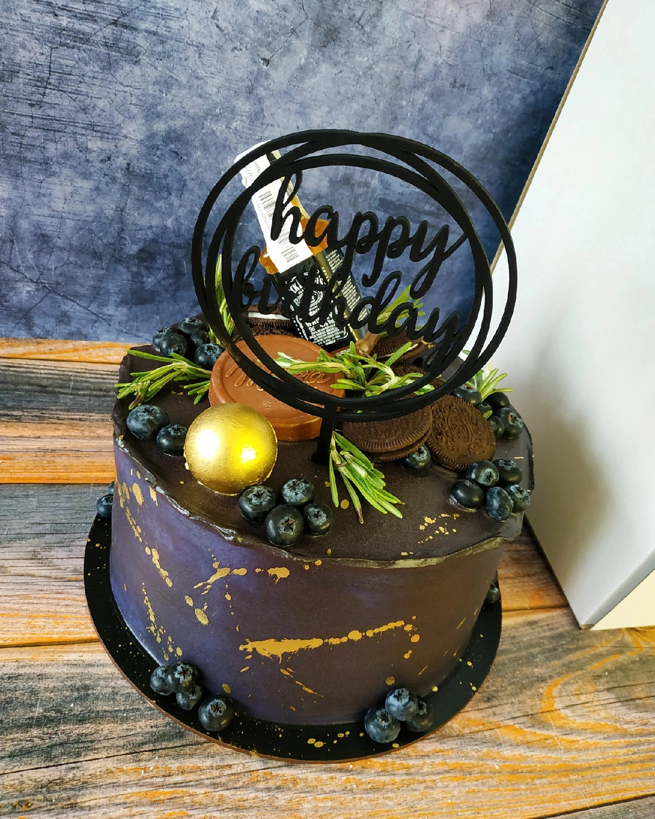 Ciemnofioletowy tort urodzinowy z dekoracjami z borówek, ciasteczek Oreo, złotą kulką, rozmarynem i topperem 'Happy Birthday' nad butelką alkoholu.
