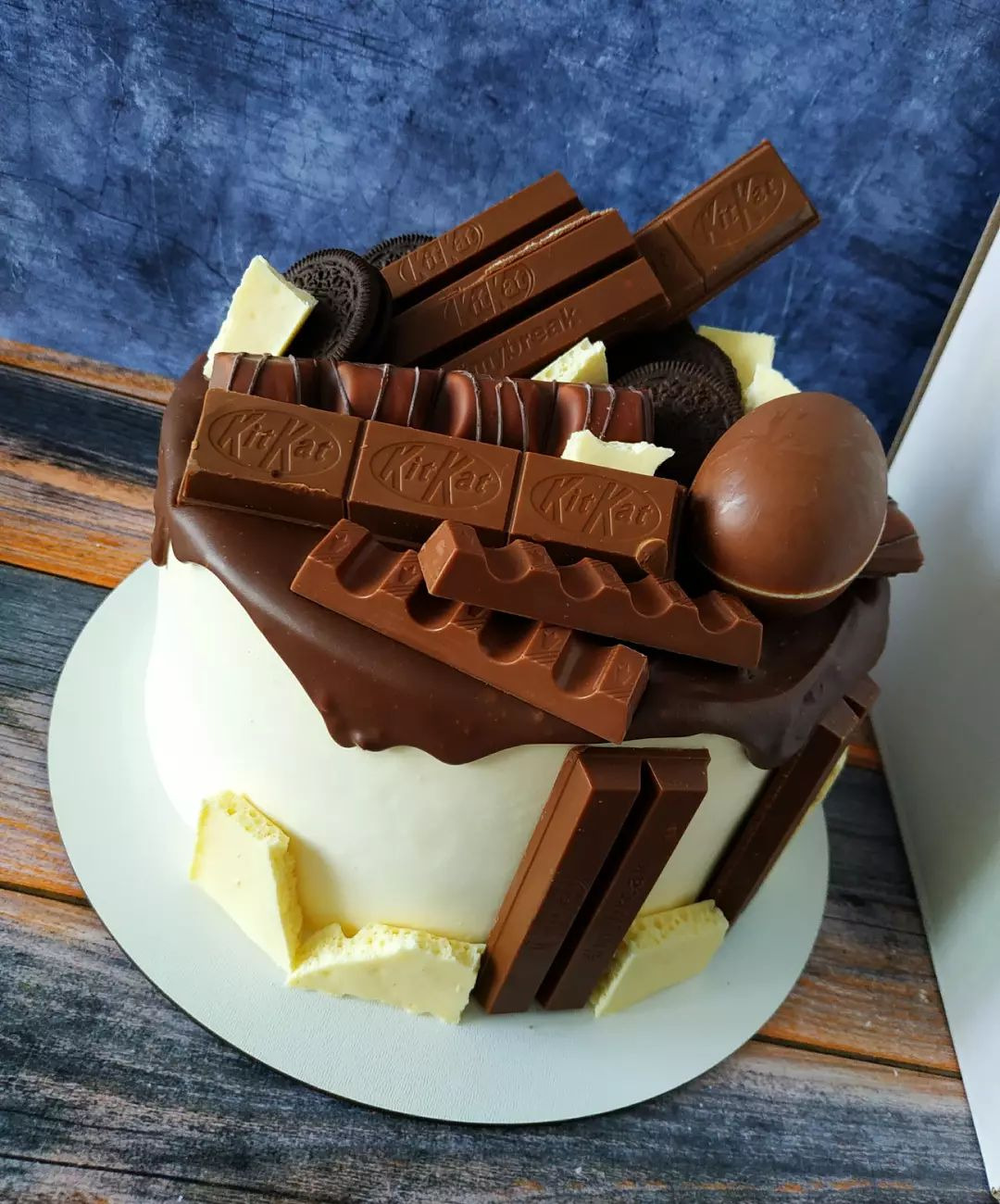 Tort z białym kremem, polewą czekoladową, udekorowany batonami KitKat, ciasteczkami Oreo i czekoladowym jajkiem na tle niebieskiej ściany.
