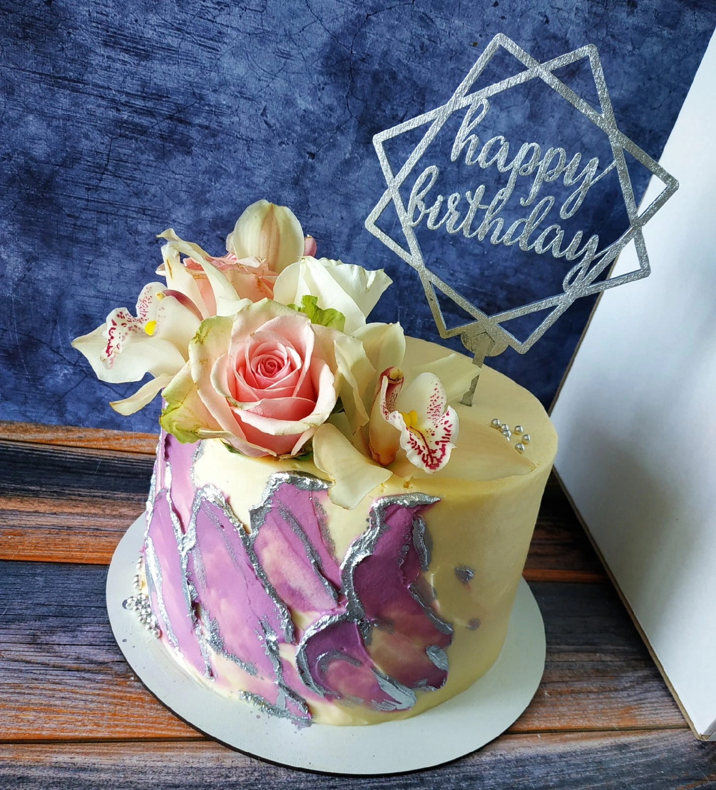 Tort urodzinowy z kremowym lukrem, dekorowany różami, orchideami i srebrnym napisem 'Happy Birthday' w geometrycznej ramce.