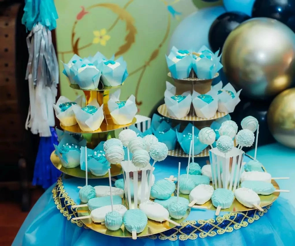 Dekoracyjny stół z babeczkami w błękitnych papilotkach, cake pops i ciasteczkami na patyku w odcieniach bieli i błękitu, ustawiony na złotym paterze z niebieską dekoracją, w tle balony i dekoracje.
