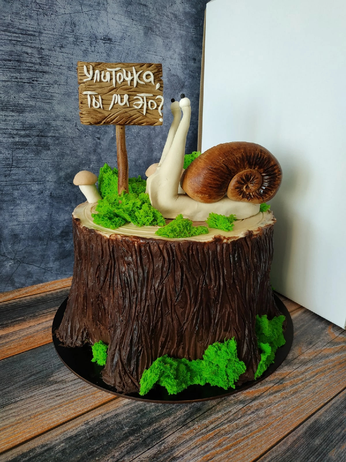 Tort w kształcie pnia drzewa z czekoladową korą, zieloną dekoracją przypominającą mech, figurką ślimaka z podniesionymi oczami i tabliczką z napisem w języku rosyjskim.