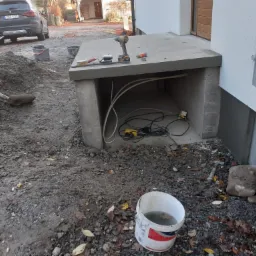 Przygotowania do prac remontowych w Łęcznej: betonowa konstrukcja z narzędziami na górze, widoczna wiertarka, szpachelka i poziomica, oraz kable i przedłużacz wewnątrz wnęki. Na ziemi wiadro z wodą...