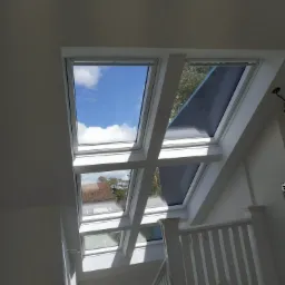 Wnętrze z białymi ścianami i drewnianą konstrukcją podtrzymującą serię okien dachowych, z których jedno ma zasuniętą roletę, a przez pozostałe widać błękitne niebo i chmury oraz fragment dachu...