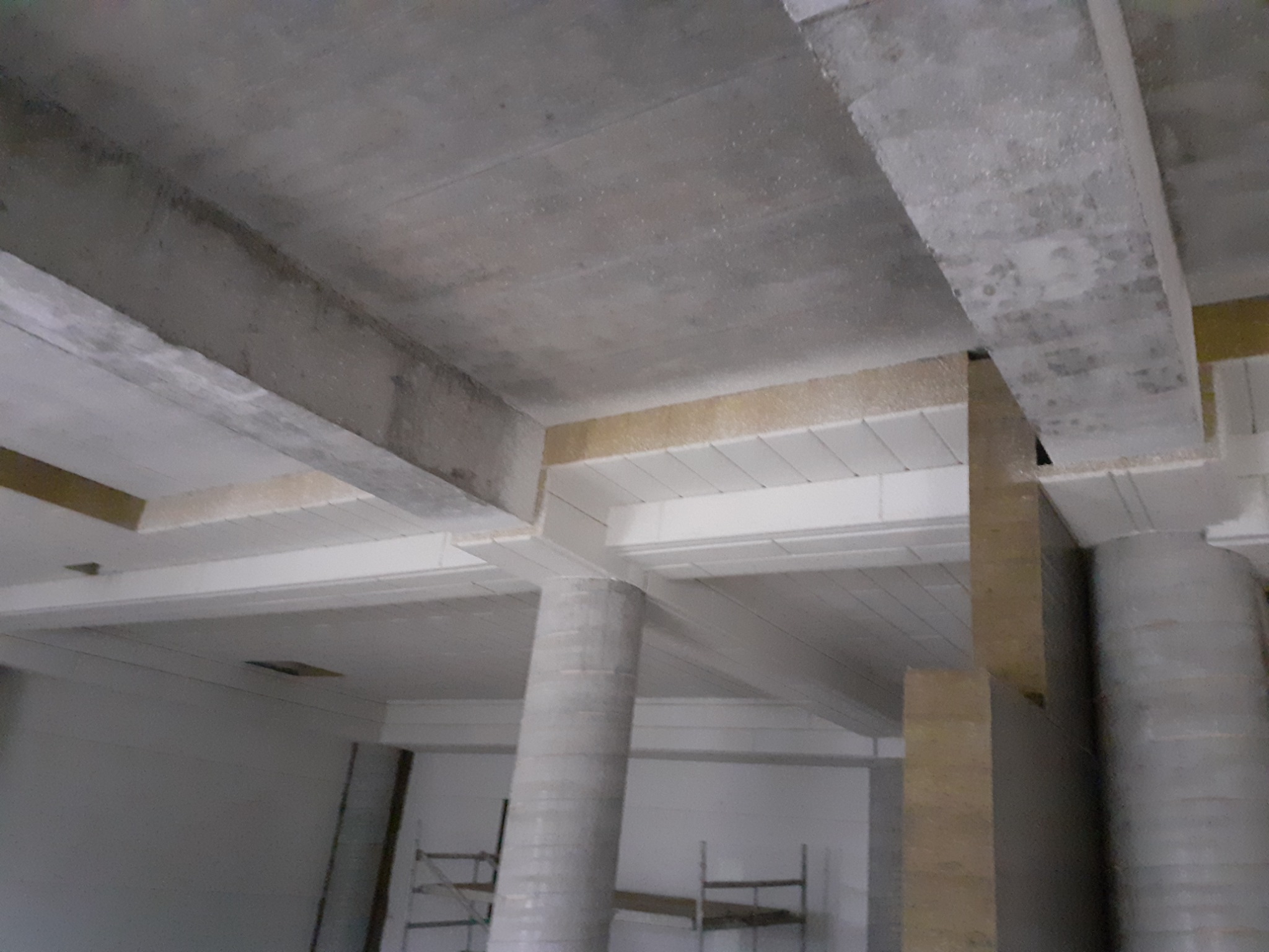 Wnętrze w trakcie prac wykończeniowych: surowy betonowy strop z widocznymi belkami, częściowo ocieplony, z kolumnami w stanie surowym i fragmentem rusztowania w tle.