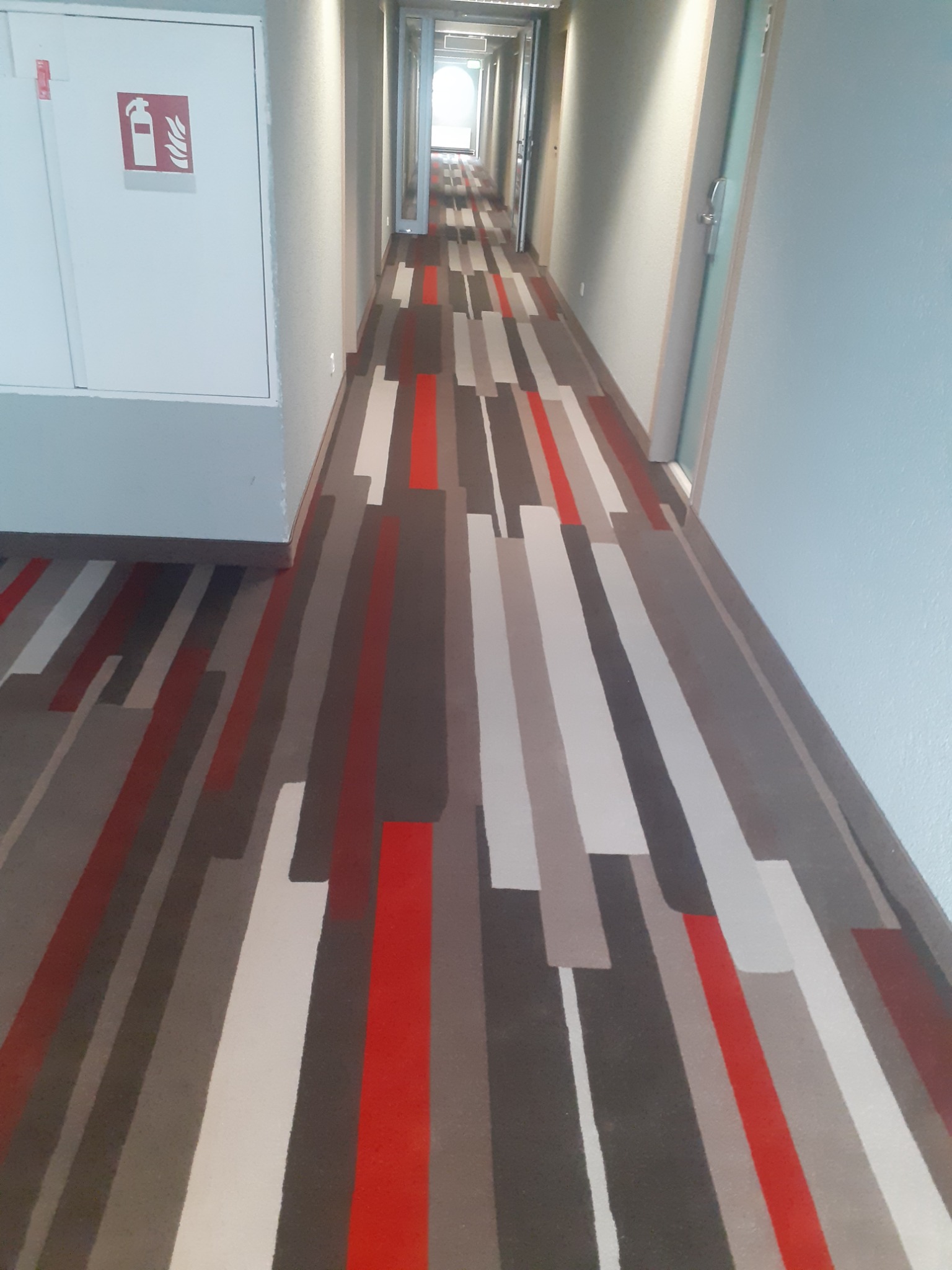 Długi korytarz hotelowy z wzorzystą wykładziną w odcieniach szarości i czerwieni, biała szafka z symbolem gaśnicy po lewej stronie.
