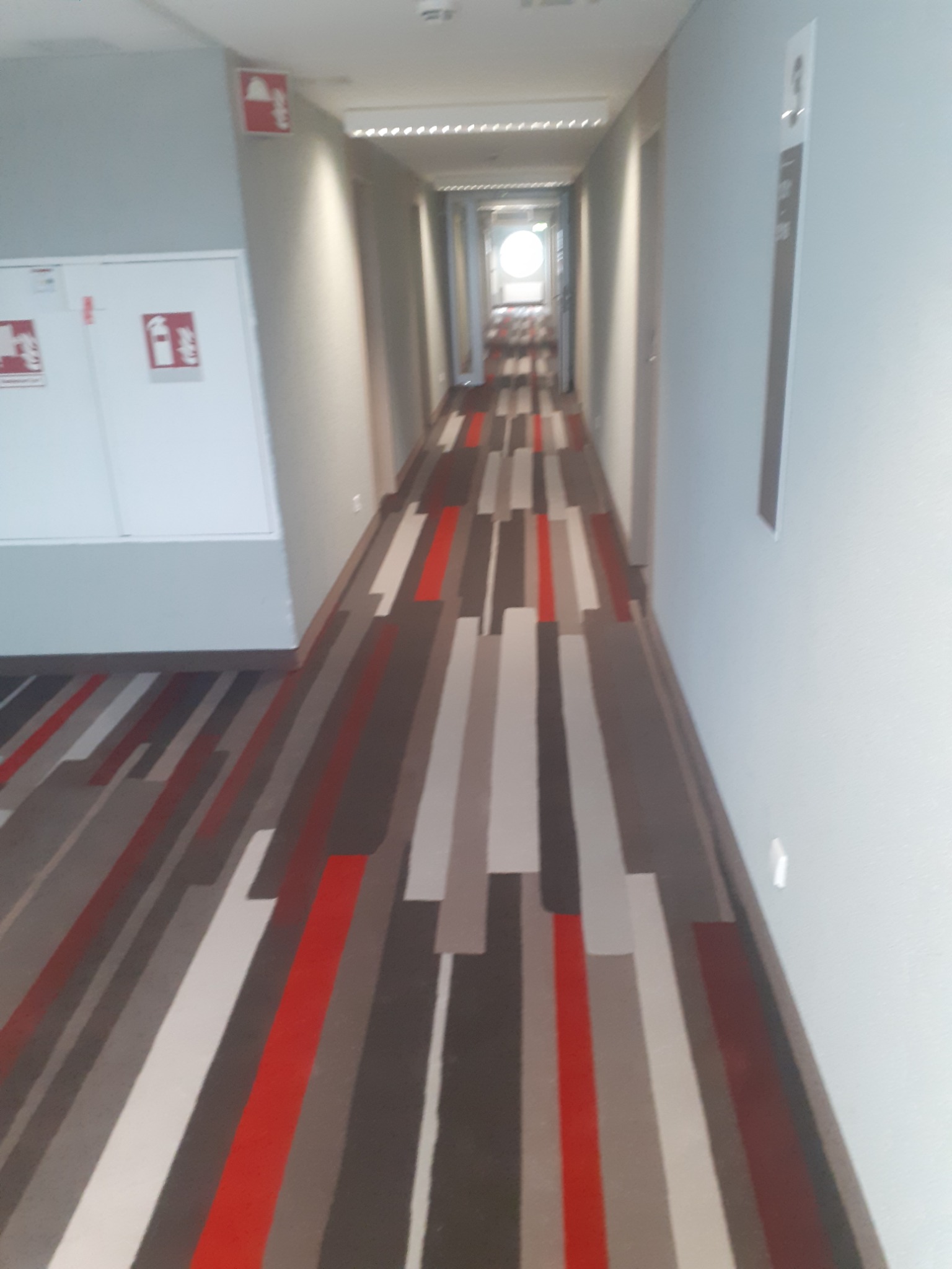 Długi korytarz hotelowy z wykładziną w geometryczne pasy (szare, białe, czerwone), białe ściany z oznaczeniami przeciwpożarowymi i numerami pokoi.