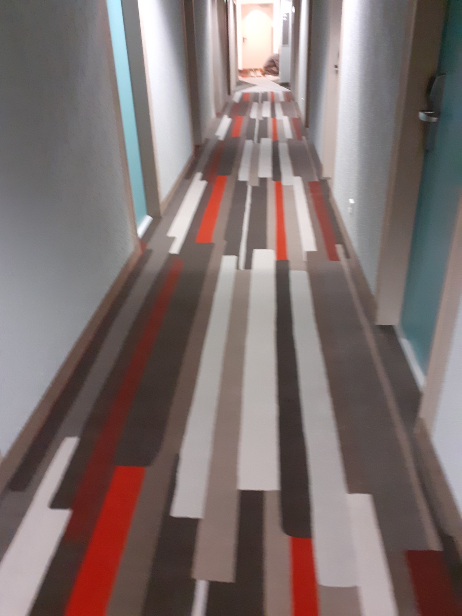 Długi korytarz hotelowy z wzorzystą wykładziną w odcieniach czerwieni, bieli i szarości, prowadzący do jasnego pomieszczenia w głębi.