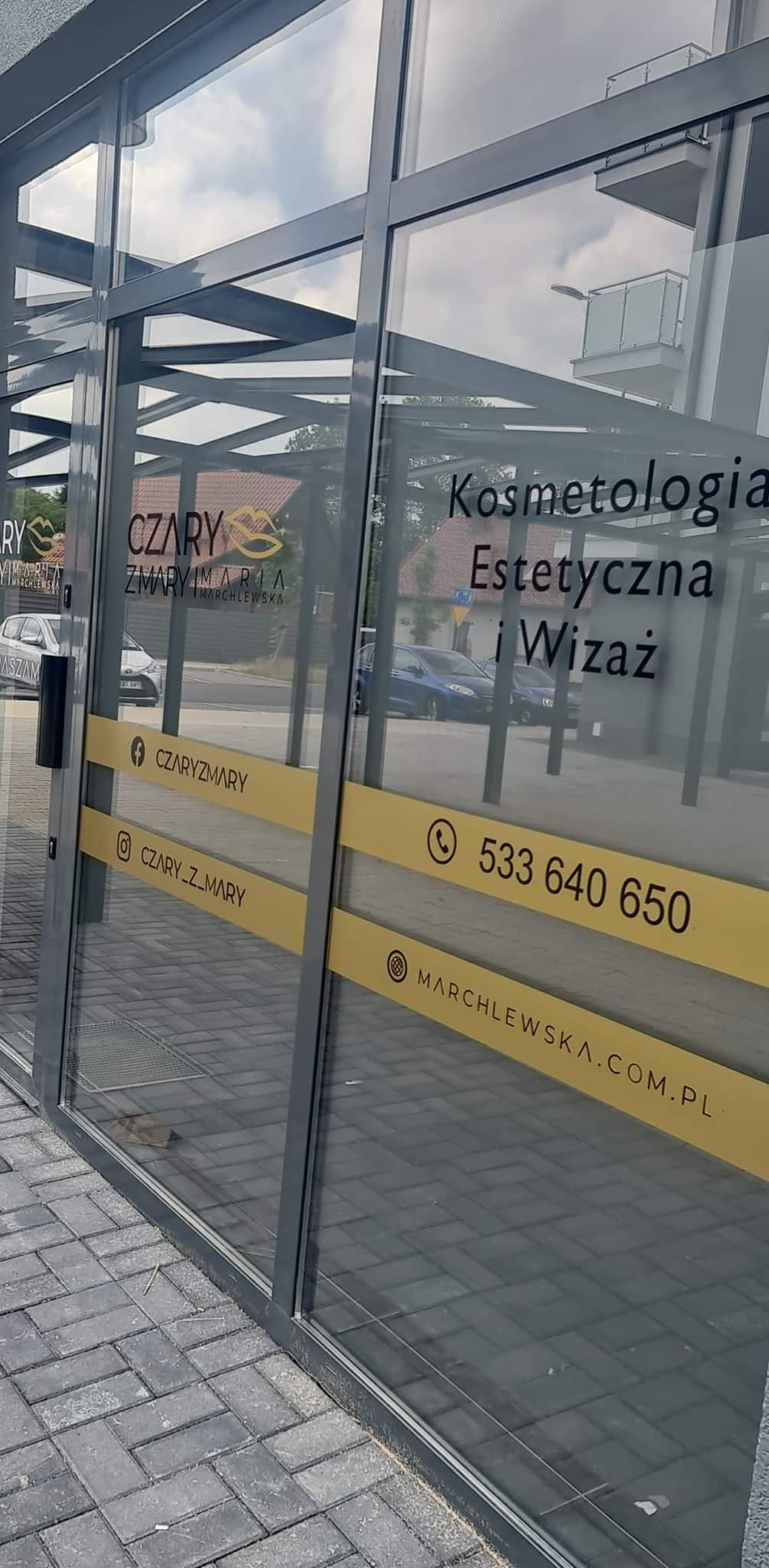 Szklane drzwi wejściowe do salonu z napisem 'Kosmetologia Estetyczna i Wizaż', logo firmy 'Czary Zmary Maria Marchlewska', danymi kontaktowymi i adresami w mediach społecznościowych na żółtych pasach.