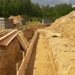 Wykop pod fundamenty z drewnianym szalunkiem, w tle ułożone bloczki betonowe i deski.