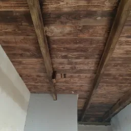 Drewniany sufit z belek, częściowo zakryty płytą kartonowo-gipsową, widoczne łączenia i niedokończone wykończenie ściany.