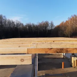 Drewniana konstrukcja stropu na wznoszonych ścianach z bloczków, widok z góry, w tle las i błękitne niebo.