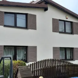 Dom jednorodzinny z białymi ścianami i brązowymi okiennicami, widok z poziomu gruntu przez częściowo otwarte drzwi samochodu dostawczego.