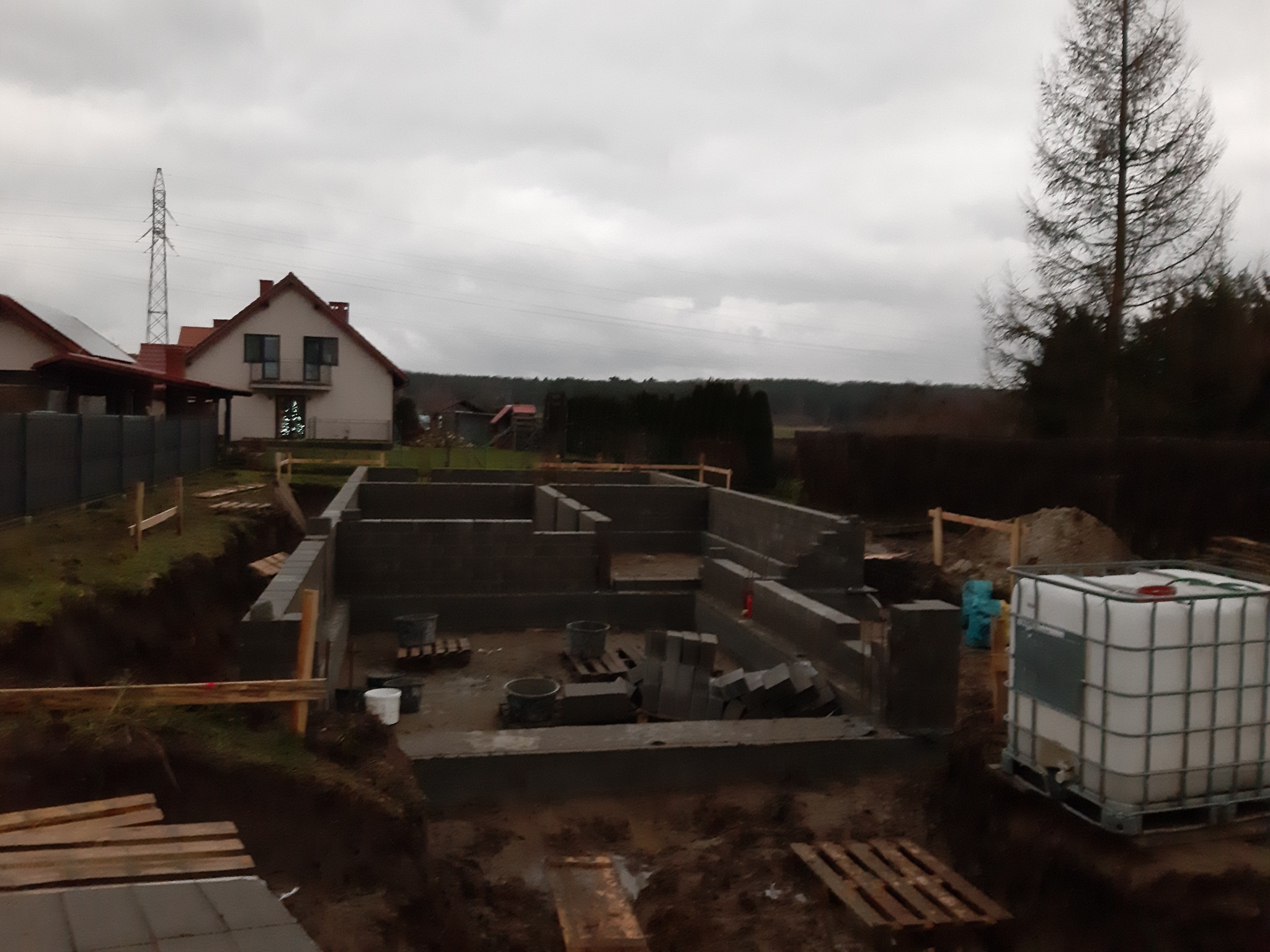 Wznoszenie ścian fundamentowych z bloczków betonowych na budowie domu jednorodzinnego, widoczne szalunki i zbiornik na wodę, pochmurne niebo w tle.
