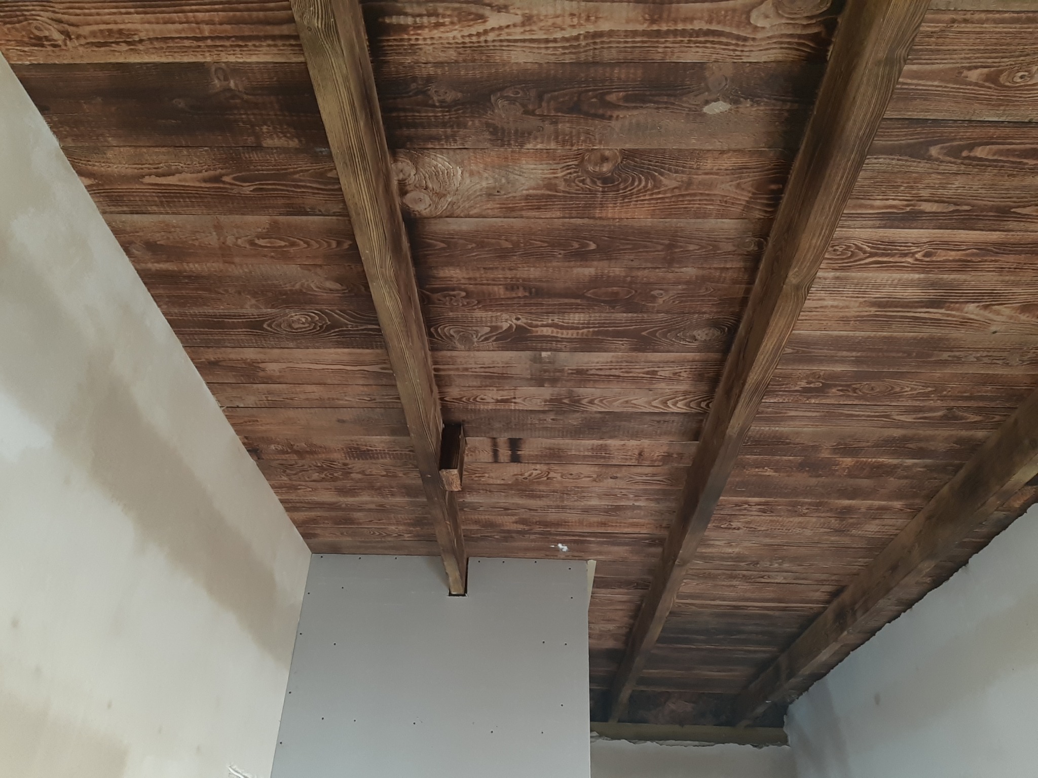 Drewniany sufit z belek, częściowo zakryty płytą kartonowo-gipsową, widoczne łączenia i niedokończone wykończenie ściany.