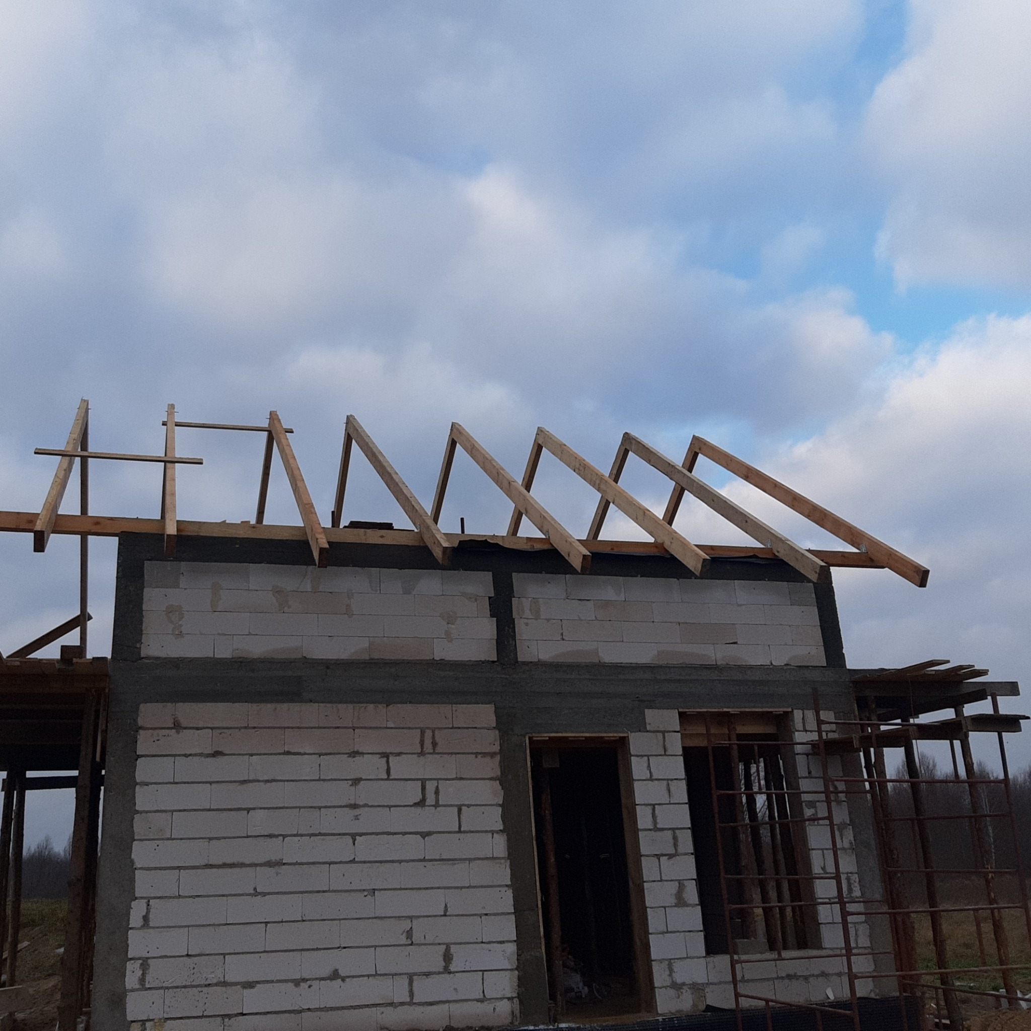 Surowy budynek z białych bloczków w trakcie budowy dachu, widoczne drewniane krokwie na tle pochmurnego nieba.