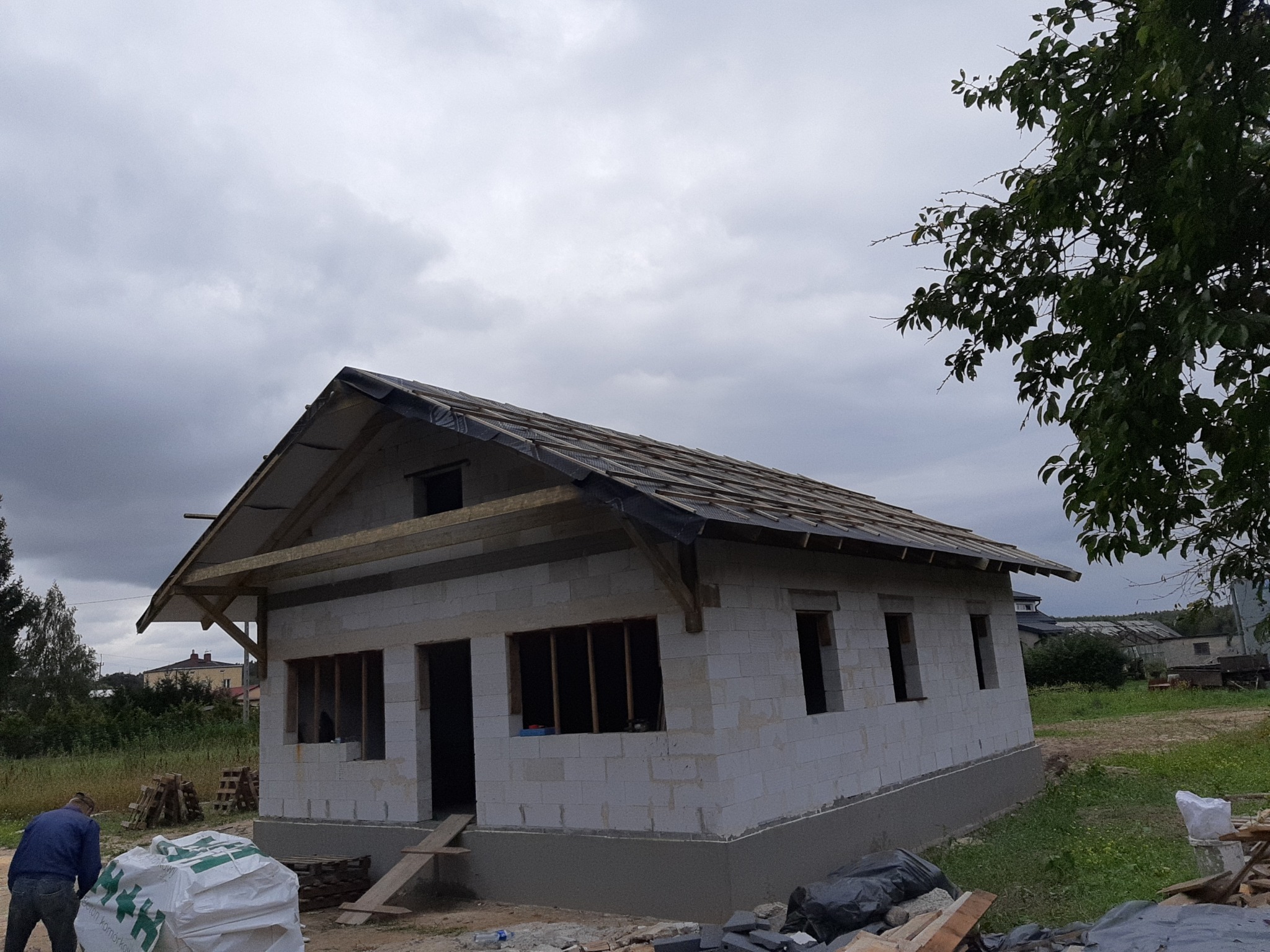 Surowy stan domu w trakcie budowy: ściany z betonu komórkowego, częściowo wykonany dach z deskami, widoczne worki z materiałami budowlanymi i pracownik w tle, pochmurne niebo.