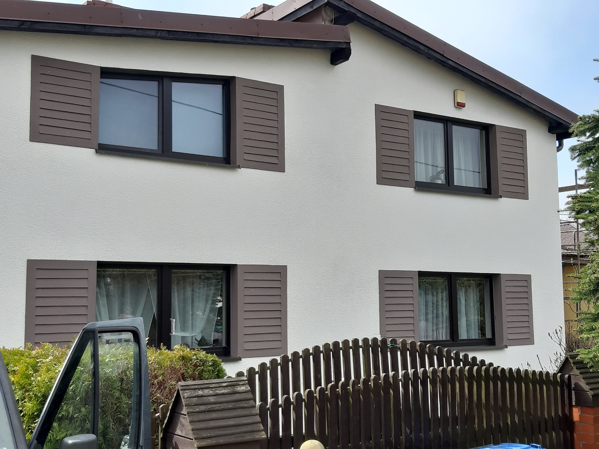 Dom jednorodzinny z białymi ścianami i brązowymi okiennicami, widok z poziomu gruntu przez częściowo otwarte drzwi samochodu dostawczego.