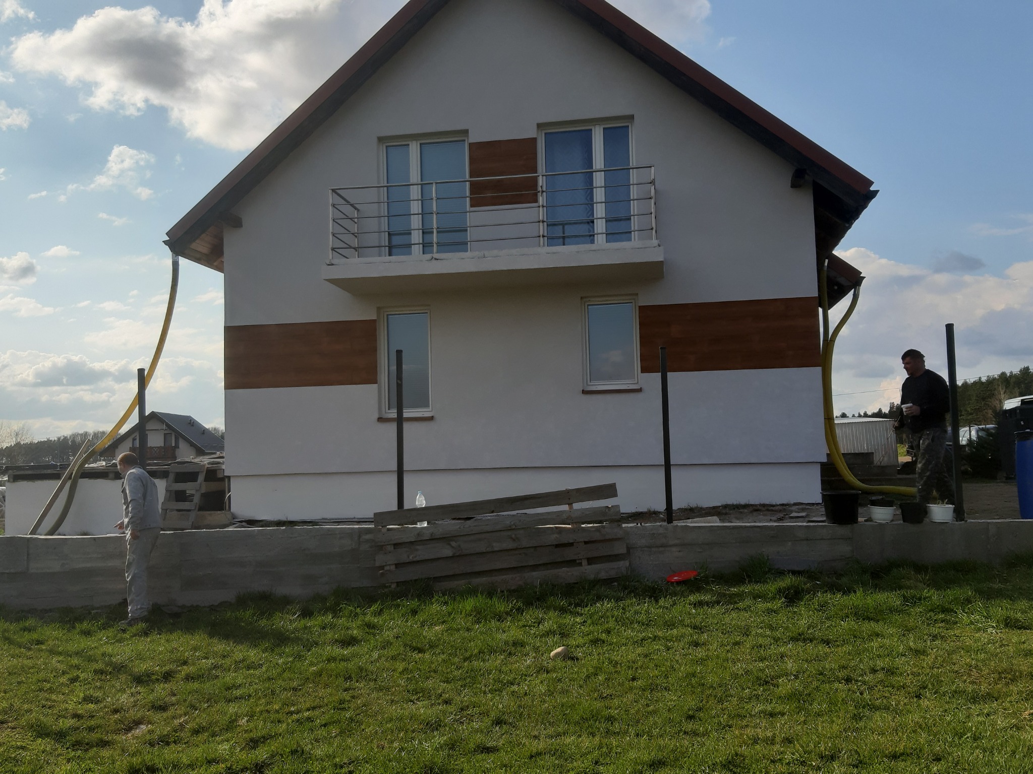 Dom jednorodzinny w trakcie natryskowego ocieplania ścian zewnętrznych, widoczne węże i pracownicy w kombinezonach ochronnych.