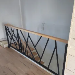 Nowoczesna balustrada schodowa z czarnymi, geometrycznymi elementami i drewnianym blatem, widok z góry na drewniane schody i podłogę, surowy stan wykończenia wnętrza.