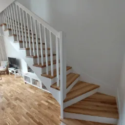 Białe schody z drewnianymi stopniami, białą balustradą i pionowymi szczeblami, widok z dołu, w tle parkiet i meble w jasnych kolorach.