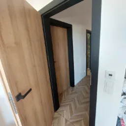 Otwarta, fornirowana skrzydło drzwiowe z czarną klamką i ościeżnicą, widok na parkiet ułożony w jodełkę w przejściu między pokojami.