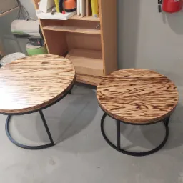 Dwa okrągłe stoliki kawowe z blatem z jasnego drewna o wyraźnym usłojeniu i czarną, metalową podstawą w kształcie łuku, ustawione w warsztacie na tle regału i ściany z gaśnicą.