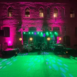 Scena muzyczna z instrumentami (perkusja, keyboard) i nagłośnieniem oświetlona zielonym i różowym światłem na tle ceglanego budynku z gotyckimi oknami.