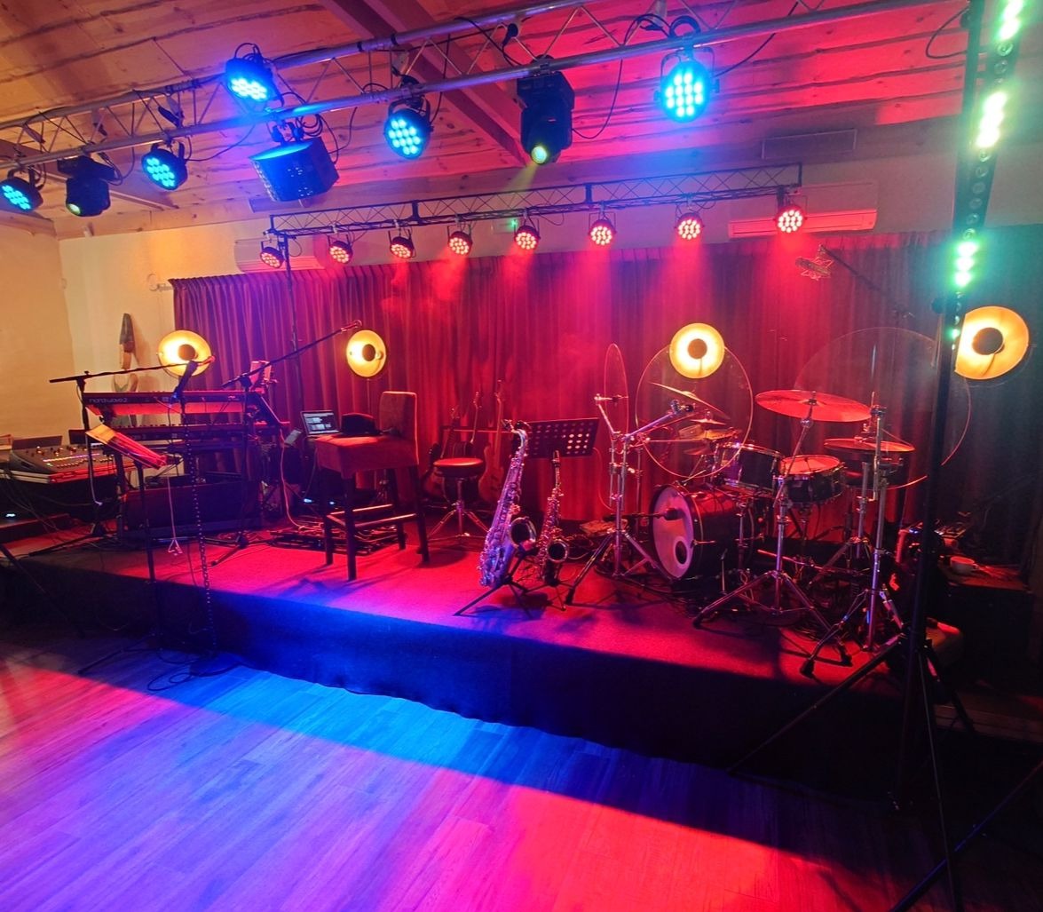 Scena z instrumentami: perkusja, saksofon, keyboard, gitary w tle, oświetlona czerwonym i niebieskim światłem. Laptop na statywie obok keyboardu.