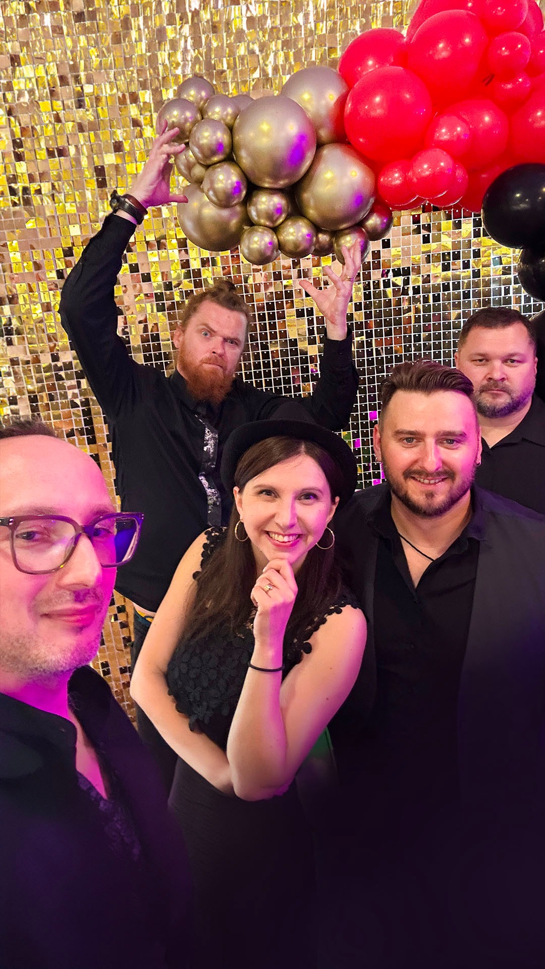 Grupa pięciu osób pozuje do zdjęcia przed błyszczącą, złotą ścianą z kwadratowych elementów, z dekoracją z czerwonych, złotych i czarnych balonów, mężczyzna z brodą trzyma balony nad głową.