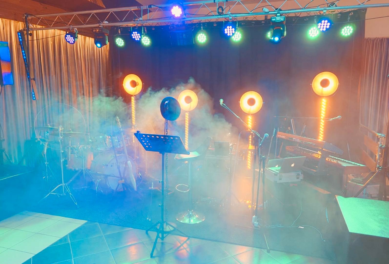 Scena przygotowana do występu: perkusja, keyboardy, mikrofony, nuty na statywie, oświetlenie sceniczne z efektem dymu.