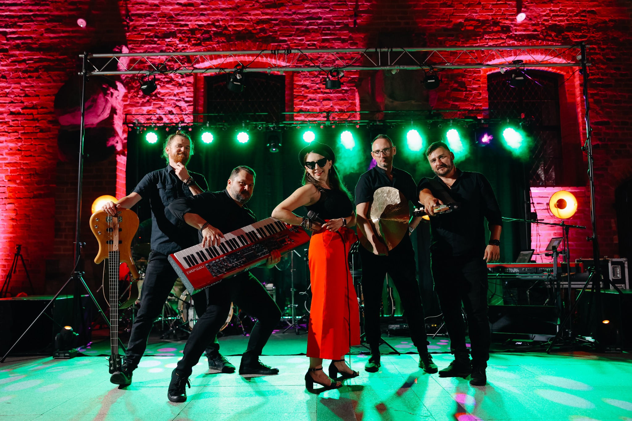 Zespół muzyczny w trakcie występu na tle czerwonej ceglanej ściany, oświetlony zielonymi reflektorami, kobieta w czerwonych spodniach trzyma mikrofon.