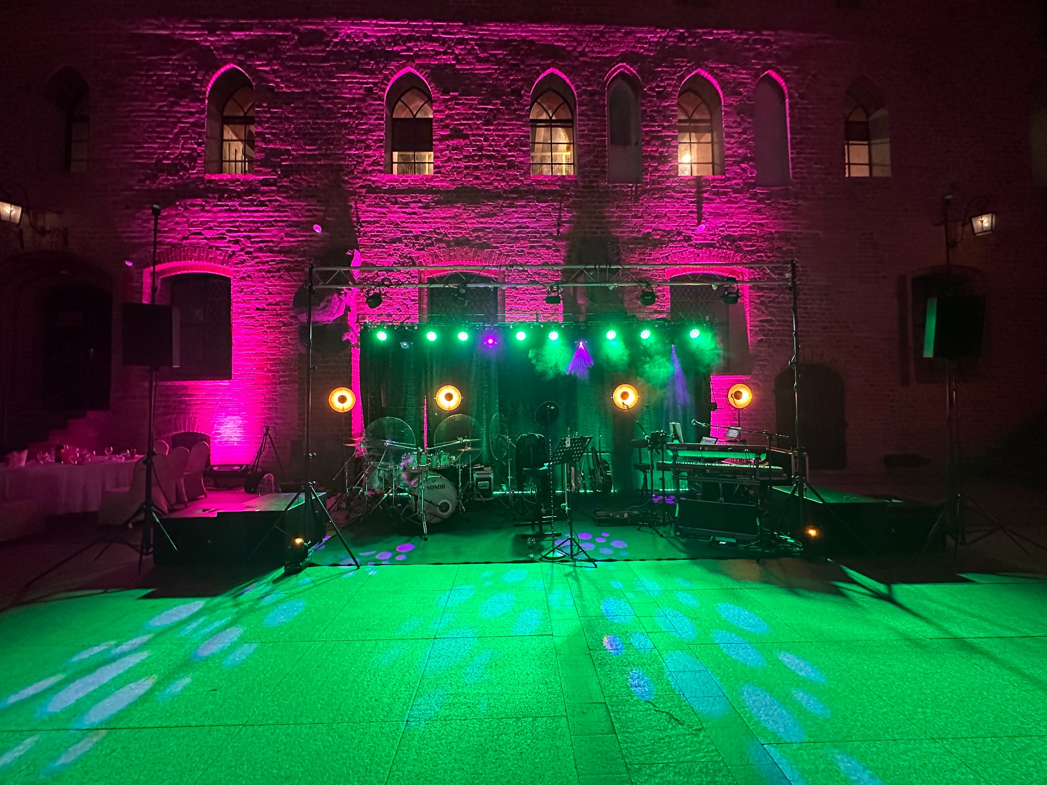 Scena muzyczna z instrumentami (perkusja, keyboard) i nagłośnieniem oświetlona zielonym i różowym światłem na tle ceglanego budynku z gotyckimi oknami.