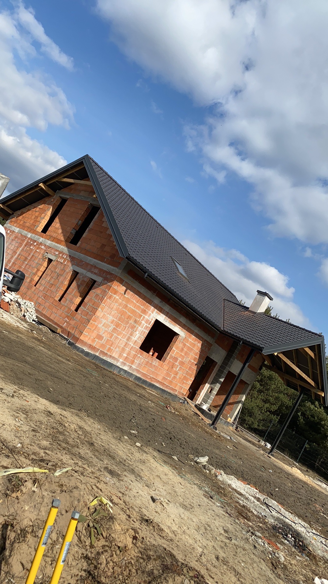 Surowy stan domu z cegły z ciemnym dachem, widoczne otwory okienne i drzwiowe, na tle błękitnego nieba z chmurami.