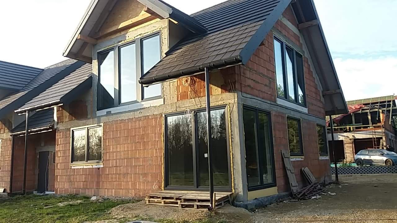 Surowy stan domu w trakcie budowy, widoczne ściany z pustaków ceramicznych, okna z czarnymi ramami, dach z ciemnej dachówki i przygotowane wejście na paletach.