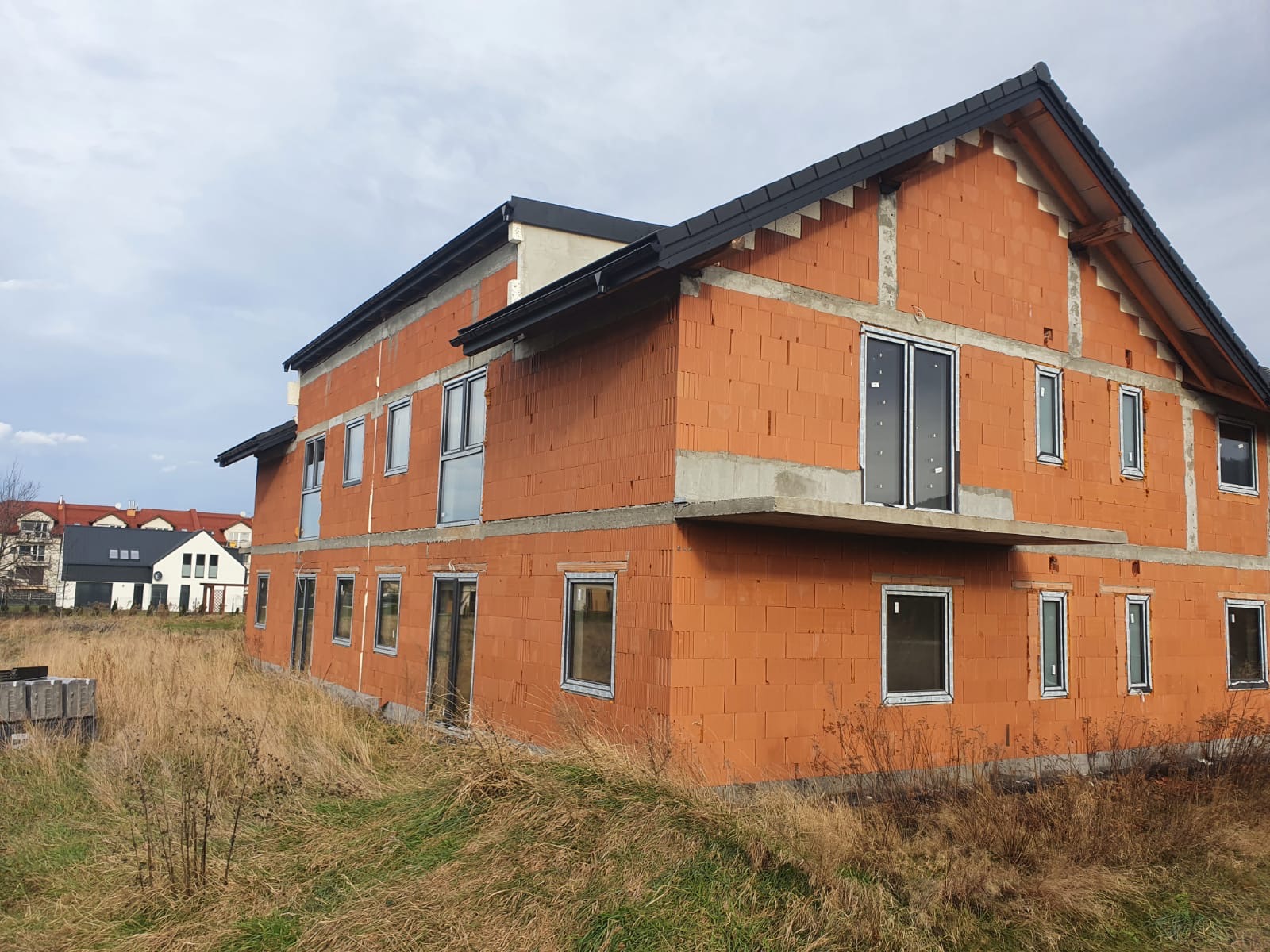 Dwukondygnacyjny dom w stanie surowym otwartym, z widocznymi oknami i drzwiami balkonowymi, wykonany z czerwonej cegły ceramicznej, z czarnym dachem i elementami betonowymi, otoczony wysoką trawą.