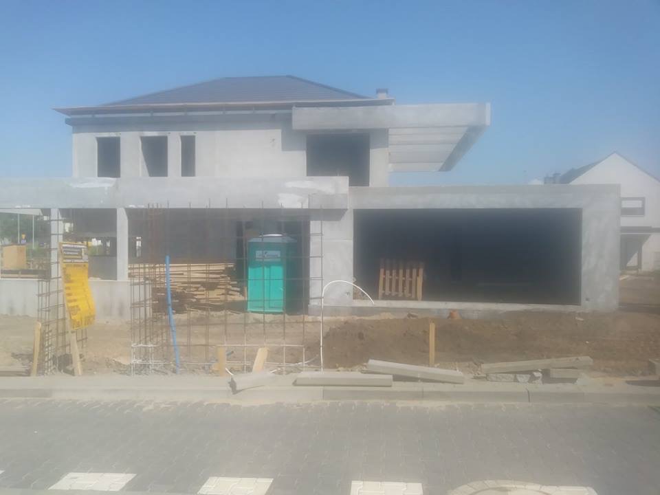 Surowy stan domu murowanego z garażem w trakcie budowy, widoczna konstrukcja żelbetowa, zbrojenie, szalunki, tymczasowa toaleta na placu budowy.