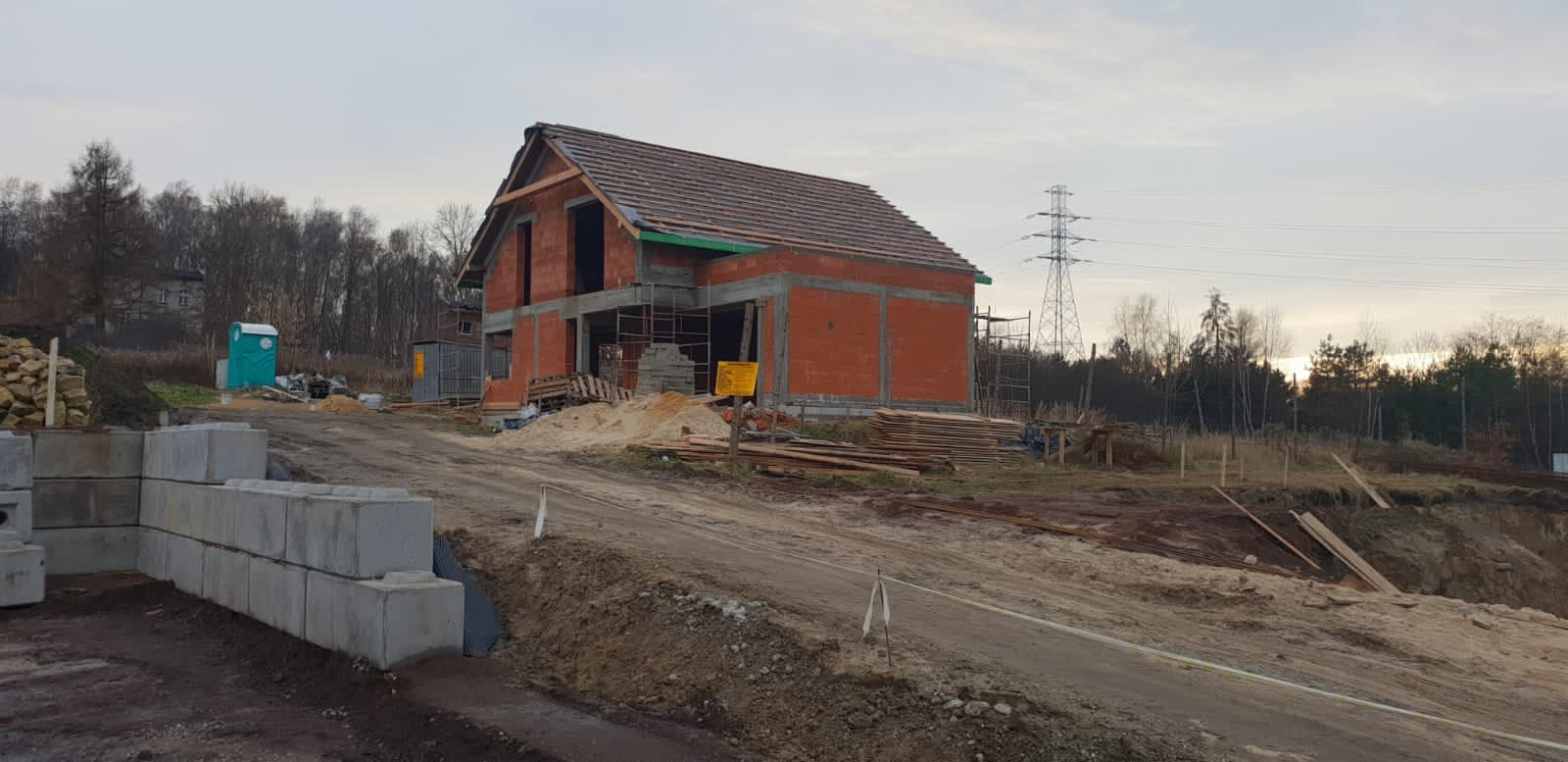 Wznoszony dwukondygnacyjny budynek z czerwonej cegły z częściowo ukończonym dachem z dachówki ceramicznej, widoczny szkielet konstrukcji i elementy zbrojeniowe, teren budowy z hałdami ziemi...