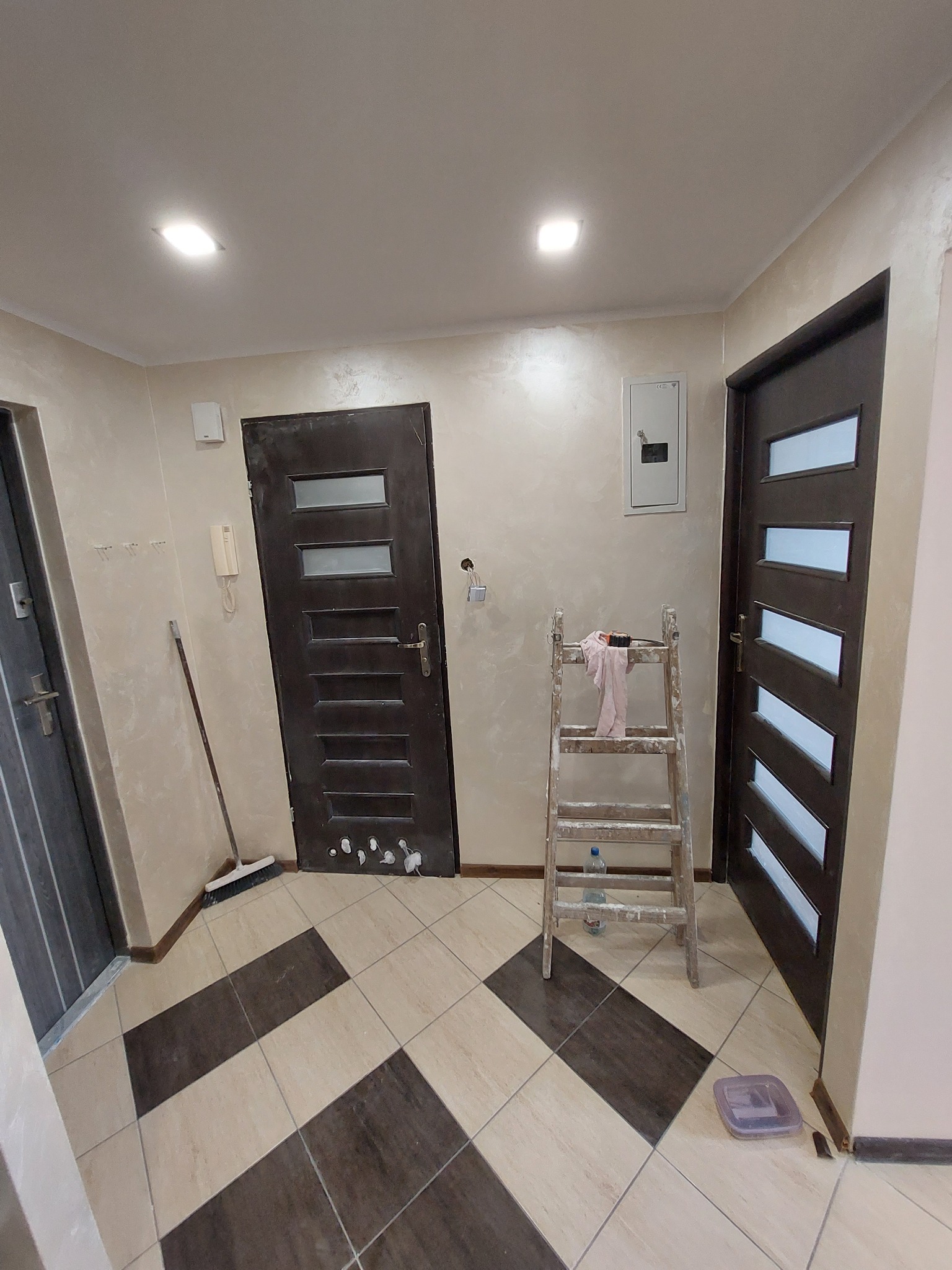 Korytarz w trakcie prac wykończeniowych: widoczna drabina malarska z akcesoriami, świeżo pomalowane ściany z efektem dekoracyjnym, dwie ciemne drzwi z przeszkleniami oraz podłoga wyłożona płytkami...