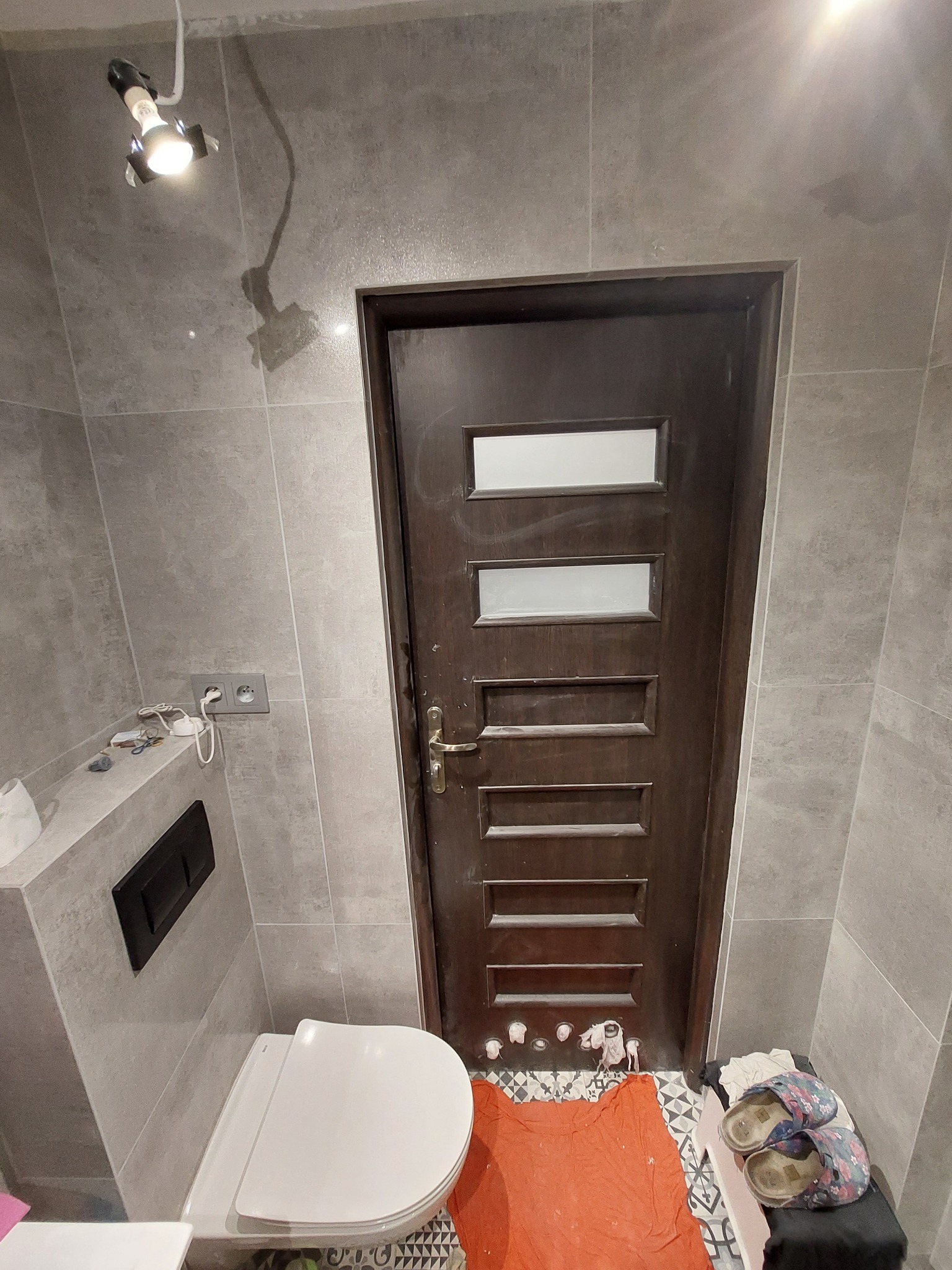 Nowoczesna łazienka w szarościach z wiszącą toaletą, ciemnymi płytkami i drewnianymi drzwiami z przeszkleniami. Na podłodze widoczne ślady prac remontowych i robocze obuwie.