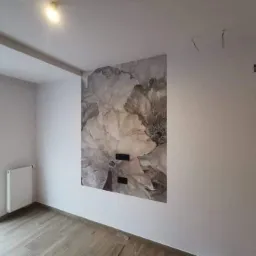 Pokój po remoncie z panelami podłogowymi i fototapetą z motywem kwiatowym na ścianie, widoczne gniazdka elektryczne.