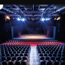 Pusta sala koncertowa z rzędami krzeseł, oświetloną sceną i reflektorami na suficie.