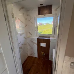 Nowoczesna toaleta z brązowymi płytkami imitującymi drewno na podłodze i częściowo na ścianie, marmurowe płytki na pozostałej części ścian, okno z widokiem na zielone pole.