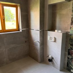 Kowalczyk-art - Łazienka w trakcie remontu: szare płytki wielkoformatowe na ścianach i podłodze, drewniane okno z widokiem na zieleń, widoczne elementy instalacji sanitarnej przed montażem, fragment dekoracyjnej...