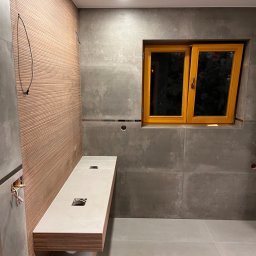 Kowalczyk-art - Łazienka w trakcie wykańczania: drewnopodobna ściana z ryflowanym wzorem, szare płytki wielkoformatowe, drewniana rama okienna, podwieszana umywalka z blatem z otworem na odpływ, widoczne kable...