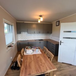 Kowalczyk-art - Wnętrze kuchni w trakcie remontu: szare szafki kuchenne z białymi kafelkami na ścianie, drewniany blat, stół z krzesłami, widoczne narzędzia i materiały budowlane na stole i pod oknem.