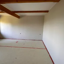 Wnętrze pokoju w trakcie wykończenia, z drewnianymi belkami stropowymi, świeżo pomalowanymi ścianami w odcieniach beżu i zabezpieczoną folią podłogą z czerwoną taśmą.