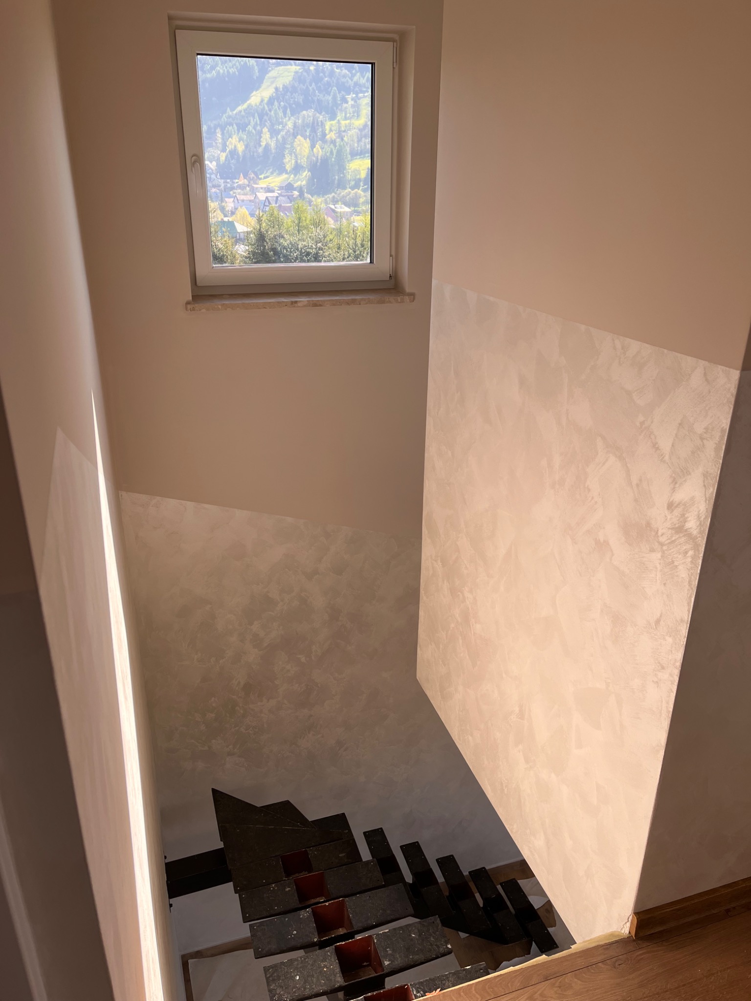 Klatka schodowa w trakcie remontu, widoczne schody tymczasowe, ściany częściowo pokryte dekoracyjną strukturą, okno z widokiem na góry.