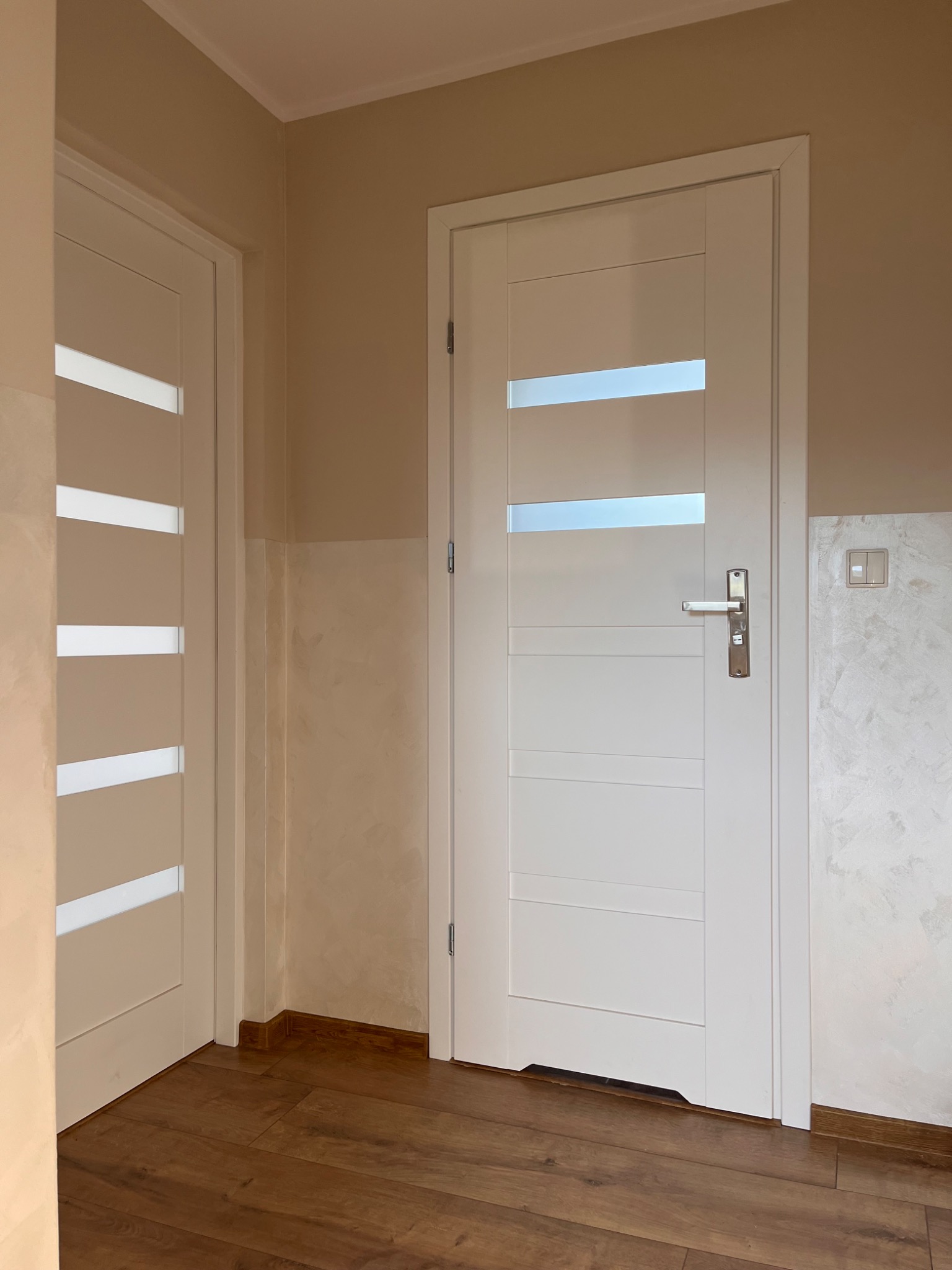 Dwie białe, nowoczesne drzwi wewnętrzne z przeszkleniami, zamontowane w pomieszczeniu z panelami podłogowymi i ścianami w odcieniach beżu z dekoracyjną fakturą.