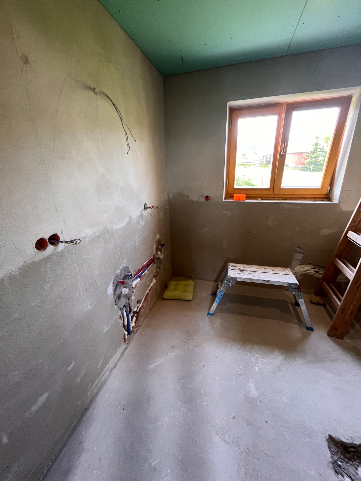 Wnętrze w trakcie prac wykończeniowych: surowa ściana z instalacją elektryczną i wodną, częściowo pokryty zieloną płytą sufit, drewniane okno z widokiem na zewnątrz, drabina i aluminiowy podest.
