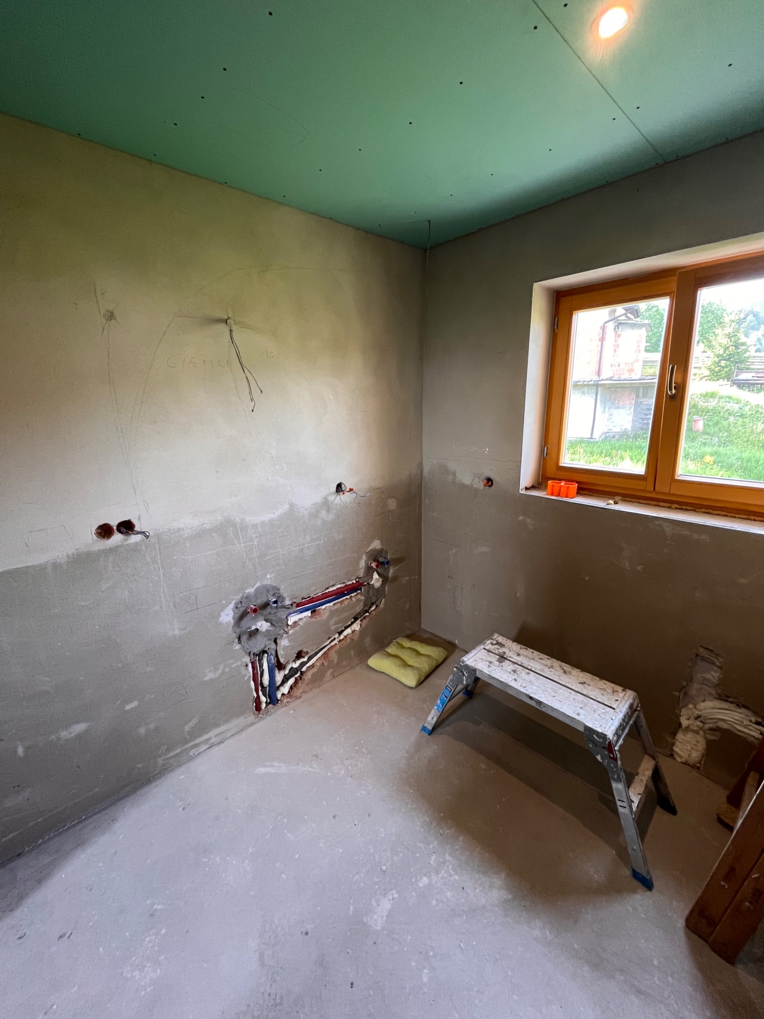 Wnętrze łazienki w trakcie remontu: surowe ściany z widocznymi instalacjami hydraulicznymi, zielony sufit z płyt kartonowo-gipsowych, okno z widokiem na zewnątrz, drabinka i poduszka na podłodze.